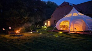 Une tente avec un feu et une chaise, installés devant un bâtiment en briques, au crépuscule.