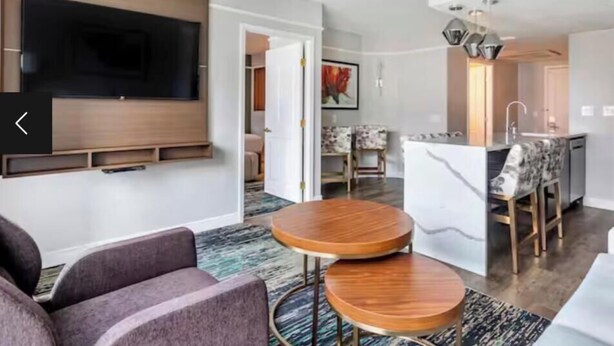 A modern living room with a flat-screen TV, a sofa, and a dining area with a kitchen in the background.