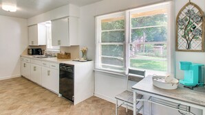 Una cocina con gabinetes blancos, lavavajillas, cafetera y ventana con persianas.