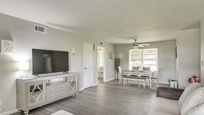 Una sala de estar con televisor, un sofá blanco, un comedor con mesa y sillas, y un ventilador de techo.