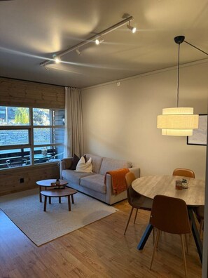 Un salon moderne comprenant un canapé, une table basse, une table à manger et des chaises. On y trouve une fenêtre avec des rideaux, un luminaire suspendu et un plancher en bois.