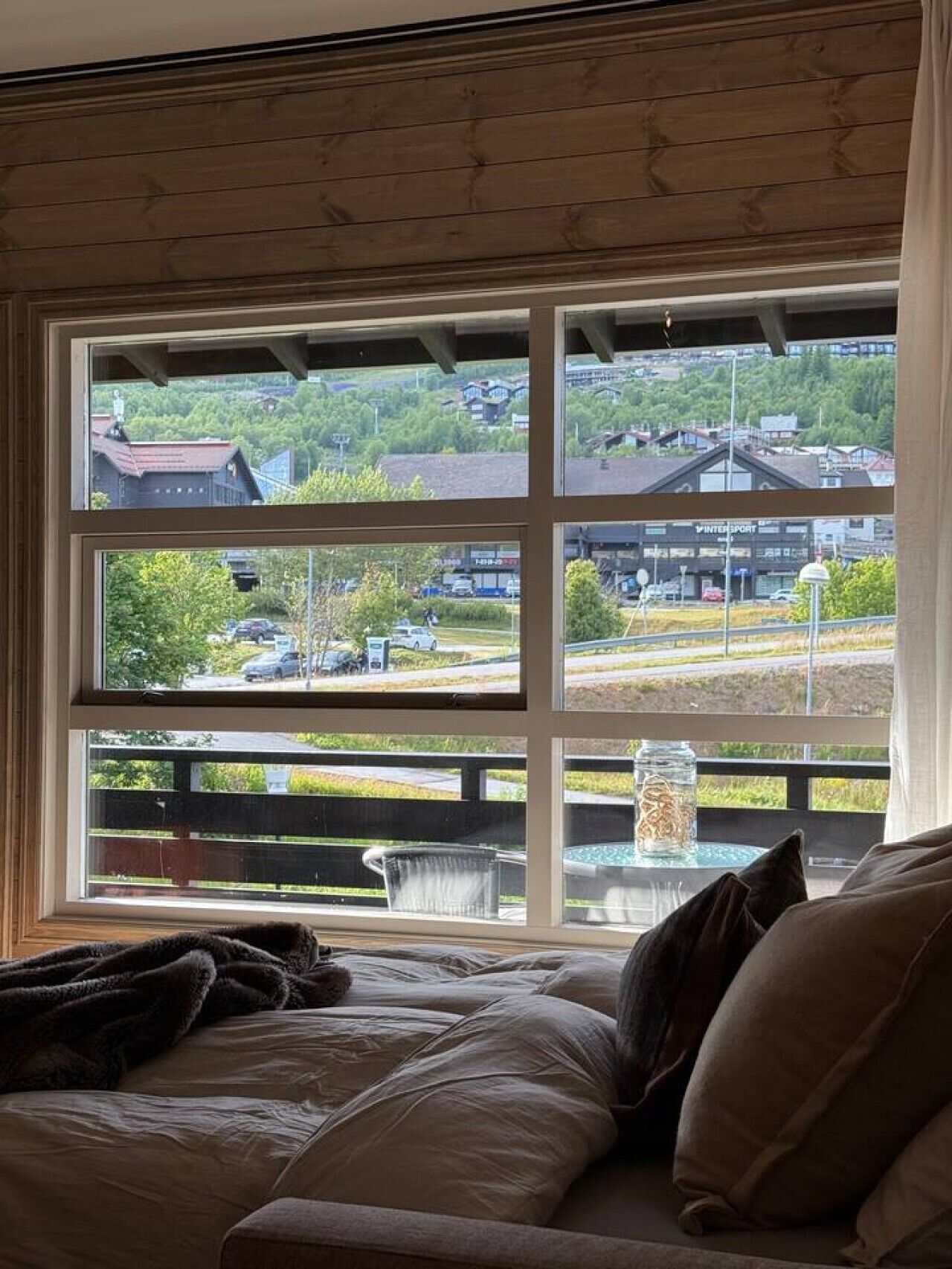 A room with a large window, a bed, and a view of a parking lot and buildings.