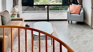 Un salon moderne avec une grande fenêtre, un canapé, un fauteuil, une petite table et un ventilateur.