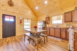 Ein Interieur einer Holzhütte mit einem Esstisch, Bänken, einem Kühlschrank, Holzausbau und einem Fenster mit Vorhängen.