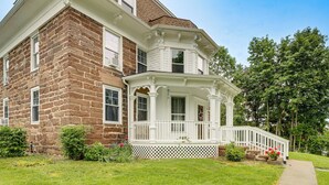 Uma casa de dois andares com uma varanda saliente, um alpendre branco com grade e um pequeno jardim frontal com grama e um caminho.