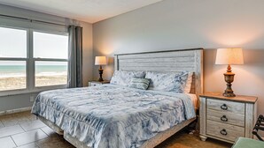 Ein Schlafzimmer mit einem Bett, zwei Nachttischlampen, einem Nachttisch, einem Sessel und Blick auf den Strand.