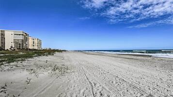 Ein weiter Sandstrand mit Fußabdrücken, ein klarer, blauer Himmel und ein Gebäude im Hintergrund.