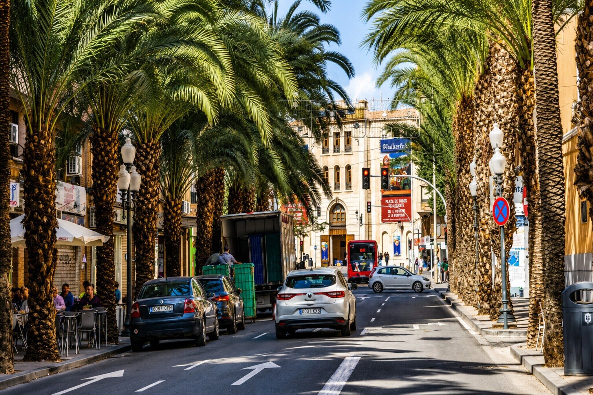 Une rue bordée de palmiers, un autobus rouge et des voitures garées le long du trottoir.