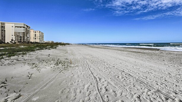 Una amplia playa de arena con huellas, un cielo azul despejado y un edificio al fondo.