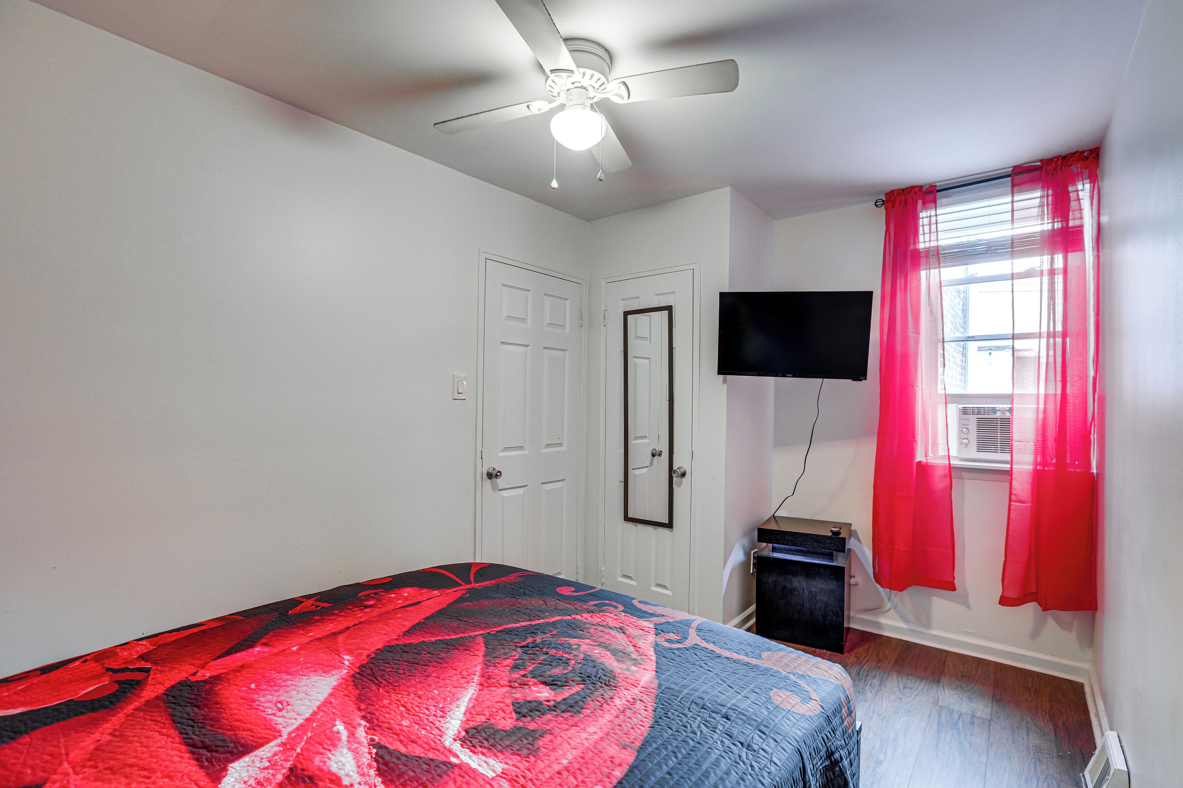 A bedroom with a bed, a ceiling fan, a TV mounted on the wall, and a window with red curtains.