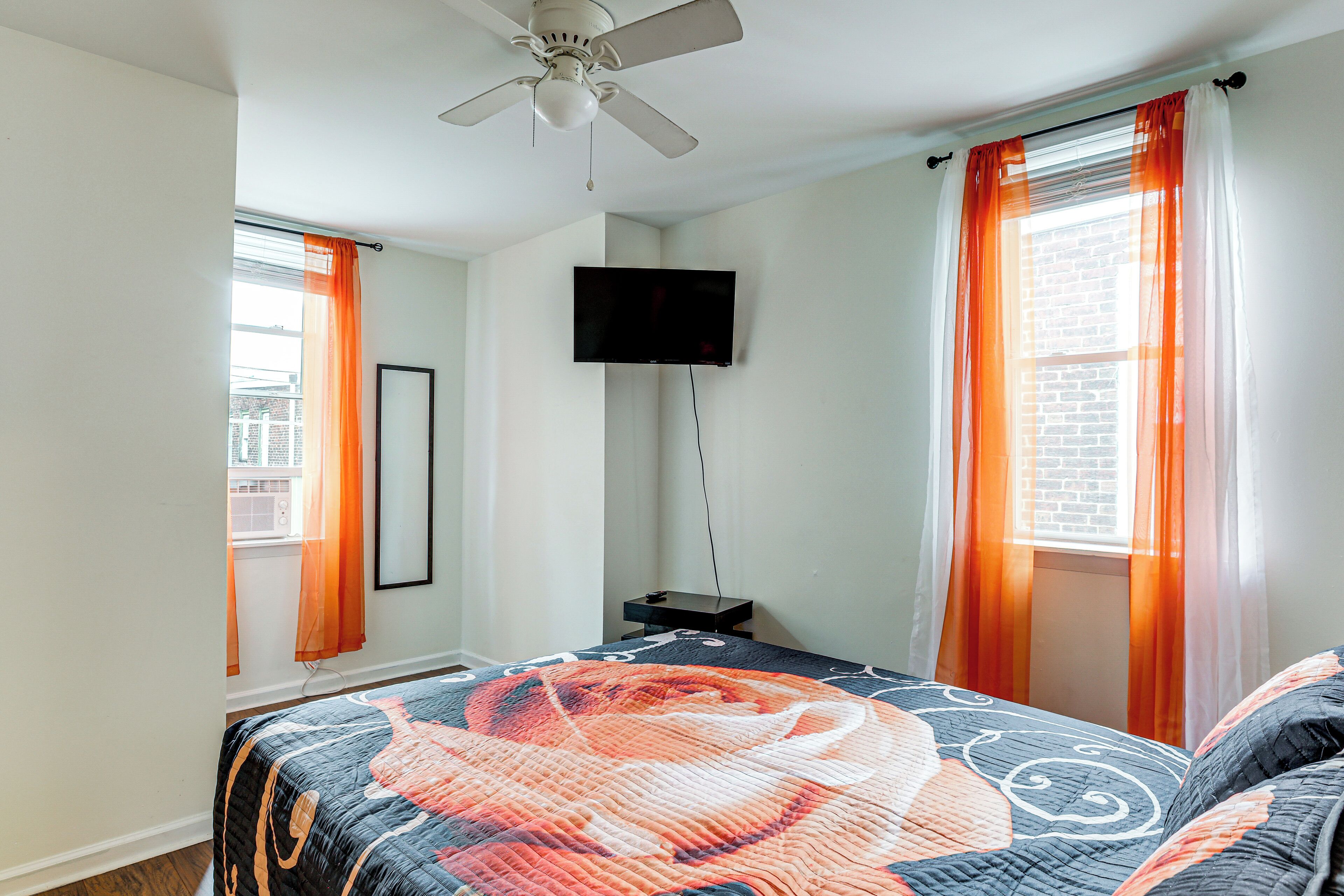 Une chambre à coucher avec un lit, un ventilateur de plafond, un téléviseur fixé au mur et une fenêtre avec des rideaux.