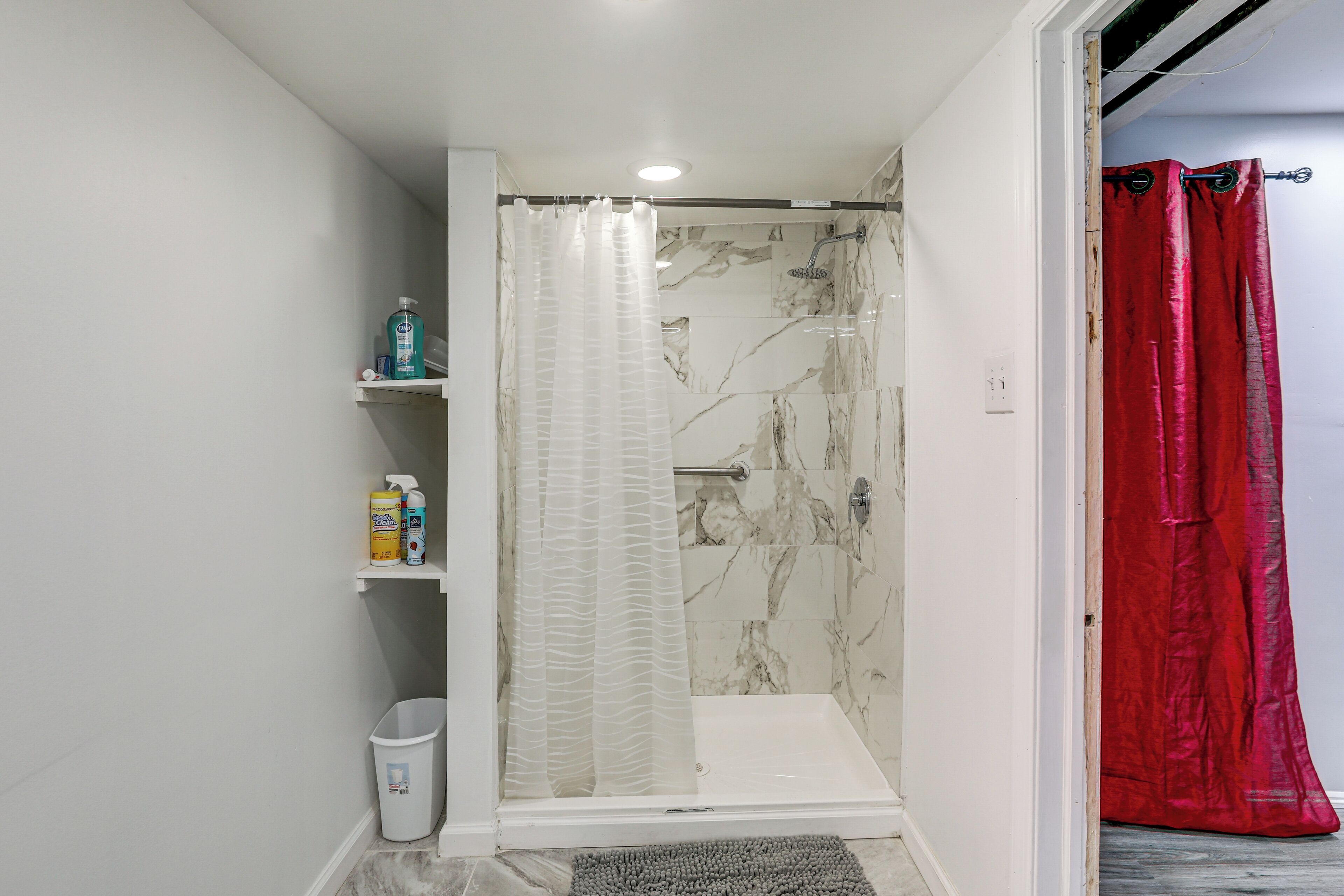 A shower with a white curtain, a red curtain, and a marble wall. There are shelves with toiletries.