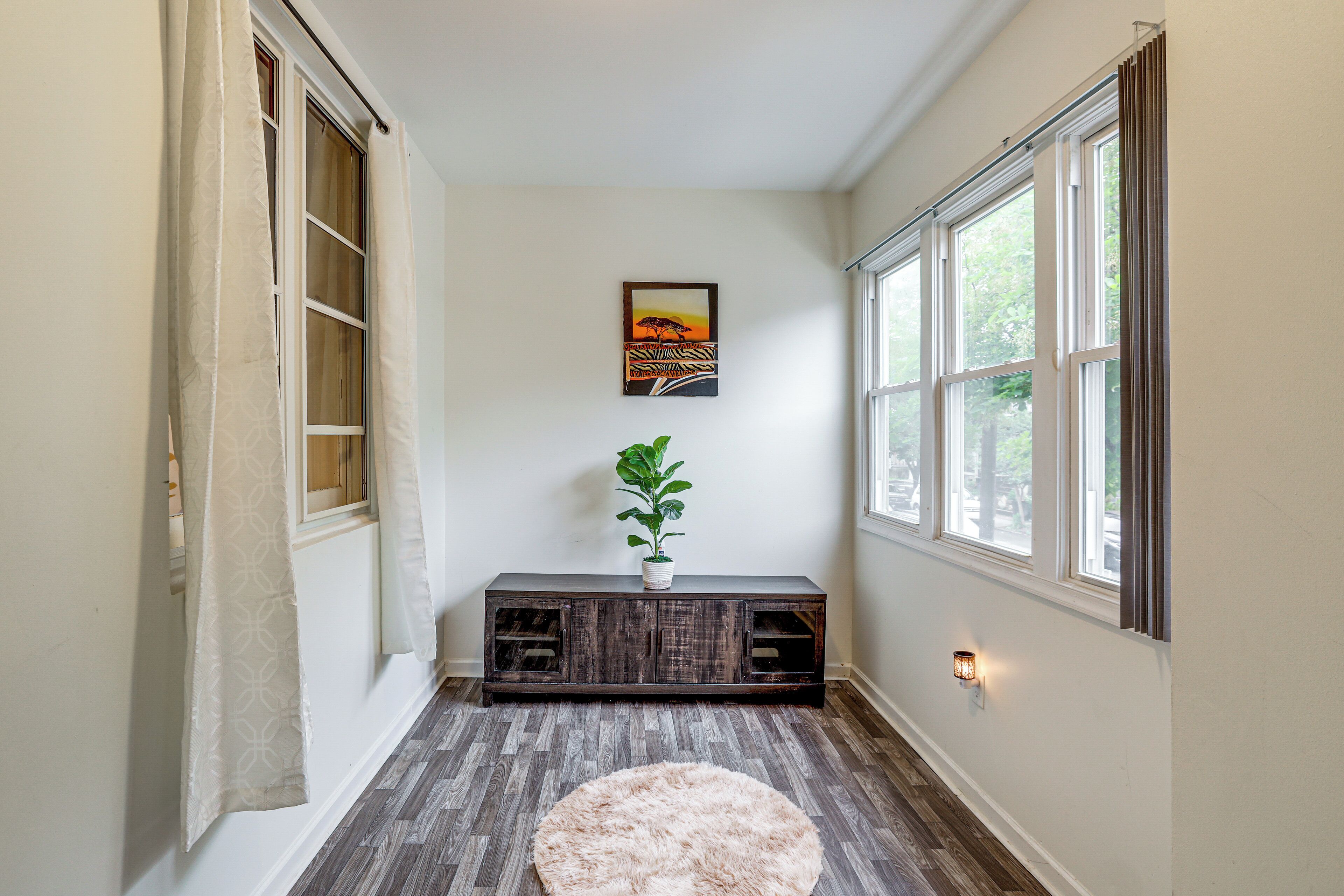 A hallway with a wooden floor, a round rug, a dark wooden bench with a plant, and a painting on the wall.