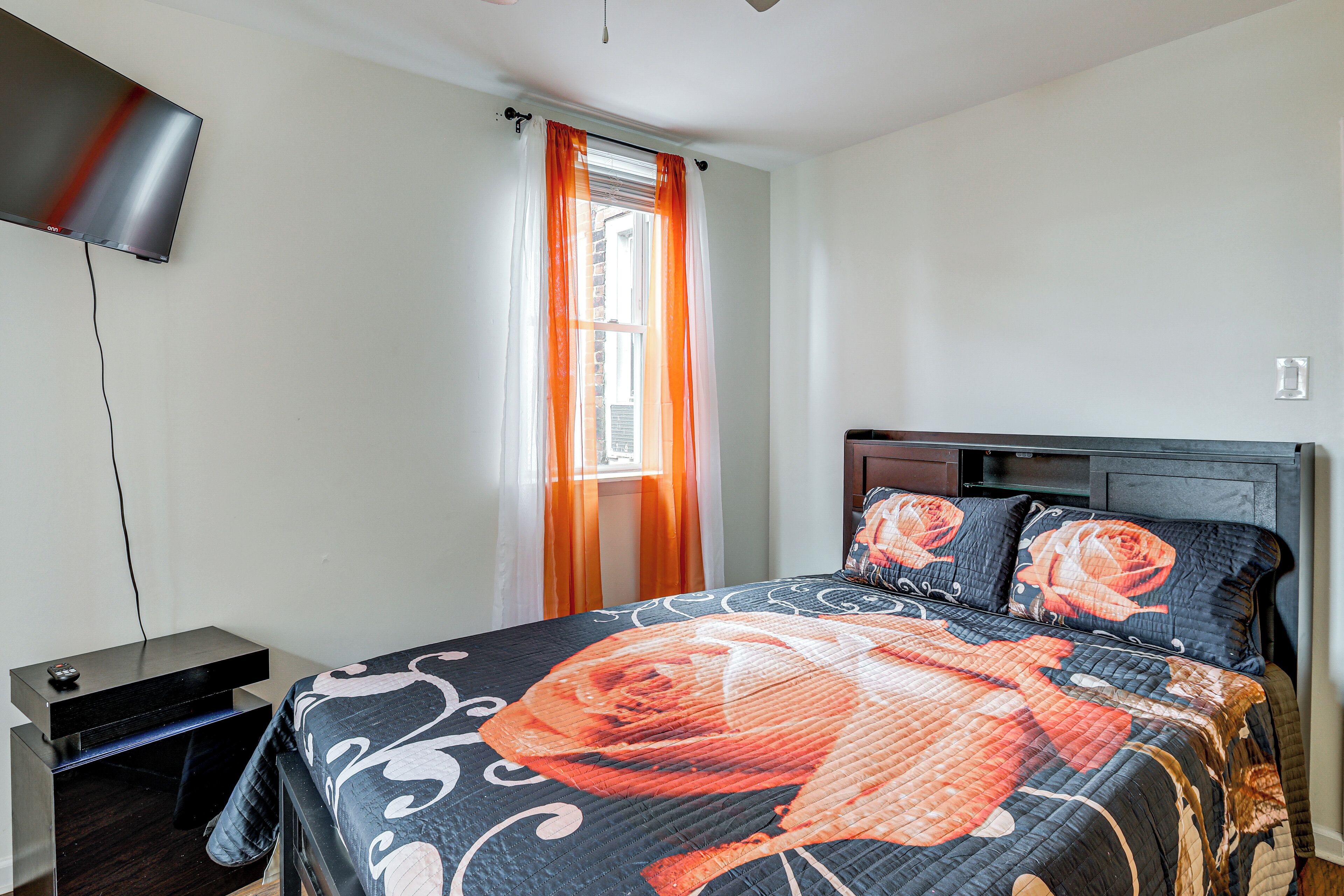 A bedroom with a bed, a TV mounted on the wall, a window with curtains, and a nightstand.