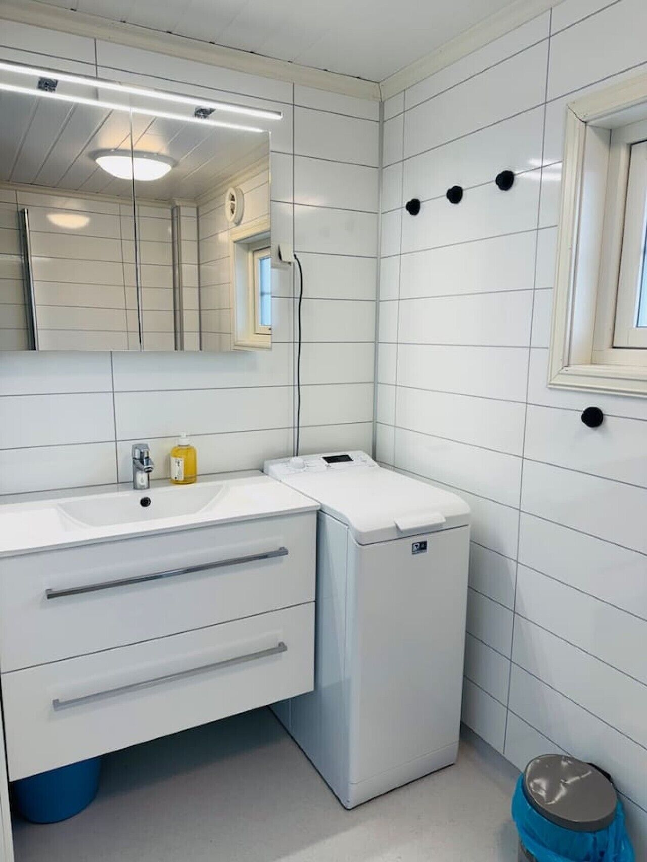 Een moderne badkamer met witte tegels, een wastafel, een spiegel, een verwarming en een prullenbak.