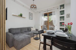 A modern living room with a grey sectional sofa, a dining table with chairs, a small round table, and a black chair. There is a shelf with plants and a picture on the wall. - Cosy Studio with Garden | Parking | Fan (Warszawa)