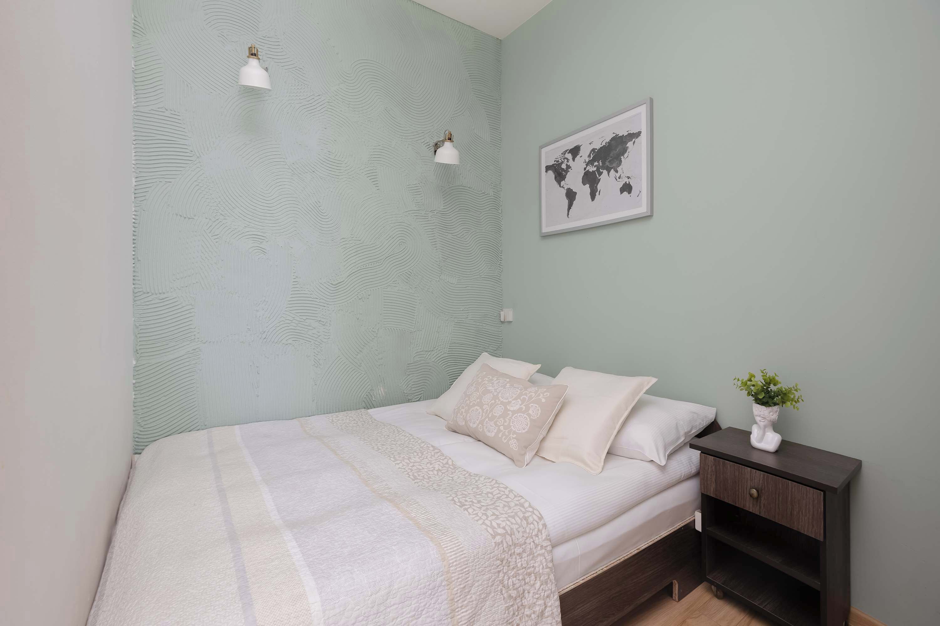 A neatly made bed with a white and beige blanket, a bedside table with a plant, and a world map on the wall.
