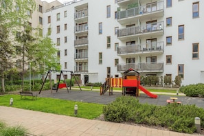 Un edificio residencial de varios pisos con balcones, un parque infantil y una banca.