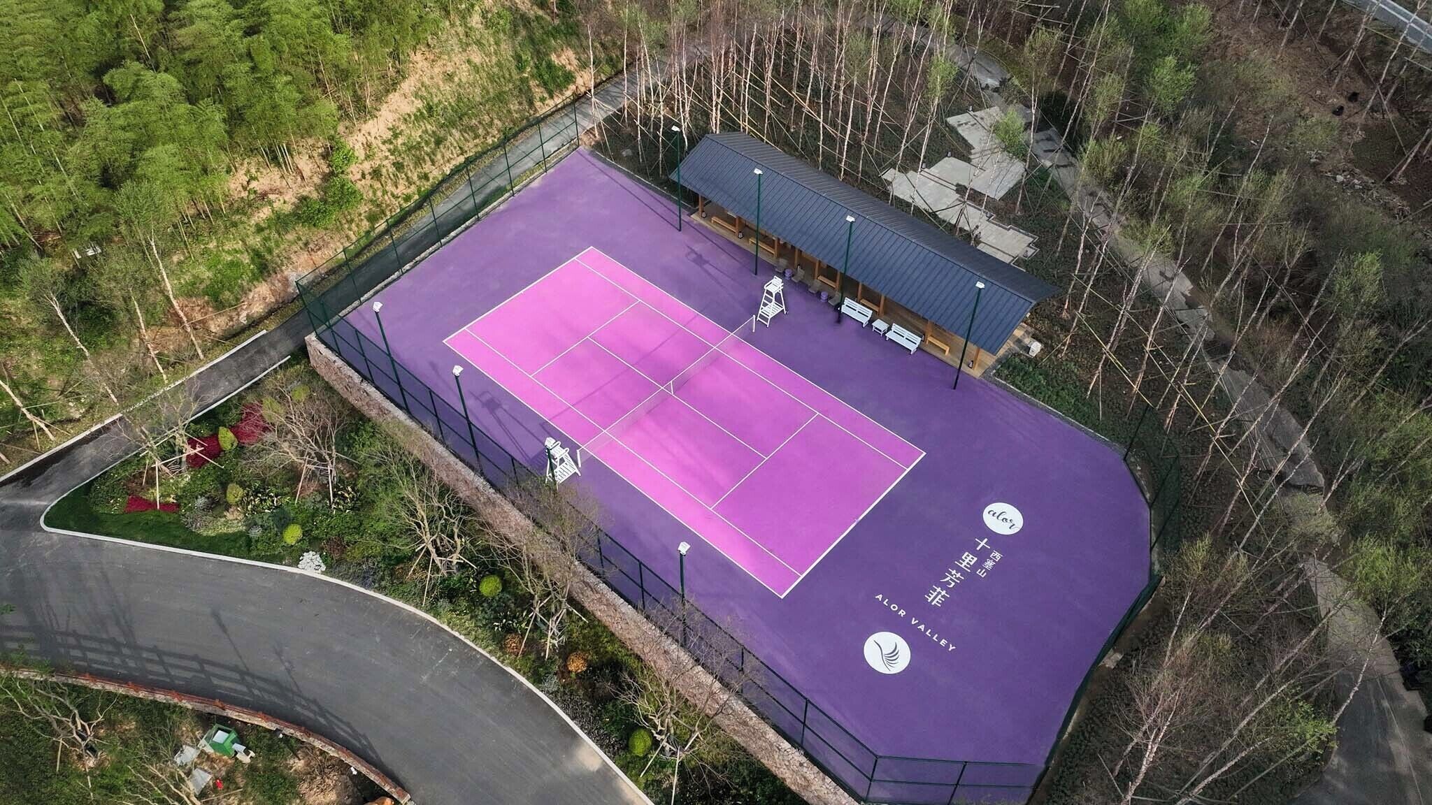 Luftaufnahme eines Tennisplatzes mit violetter Oberfläche, umgeben von Grünflächen und einer überdachten Zuschauertribüne.