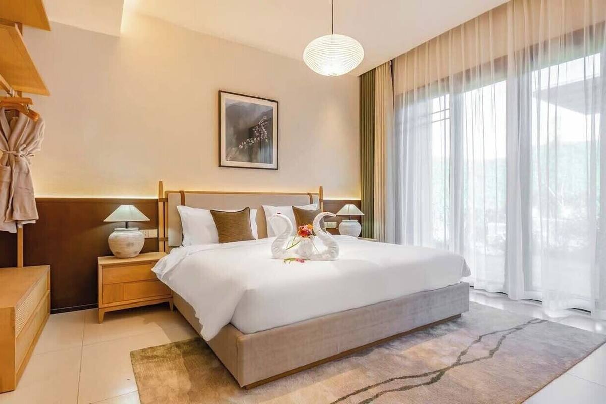 Habitación de hotel con cama, mesitas de noche, lámpara, un cuadro y un gran ventanal con cortinas.