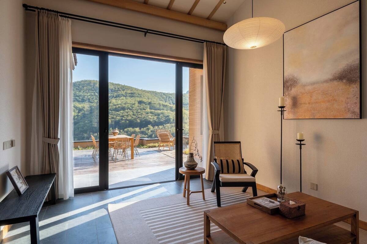 Una habitación con un amplio ventanal corredero que da a un patio exterior, una mesa de madera, una silla y un sofá.