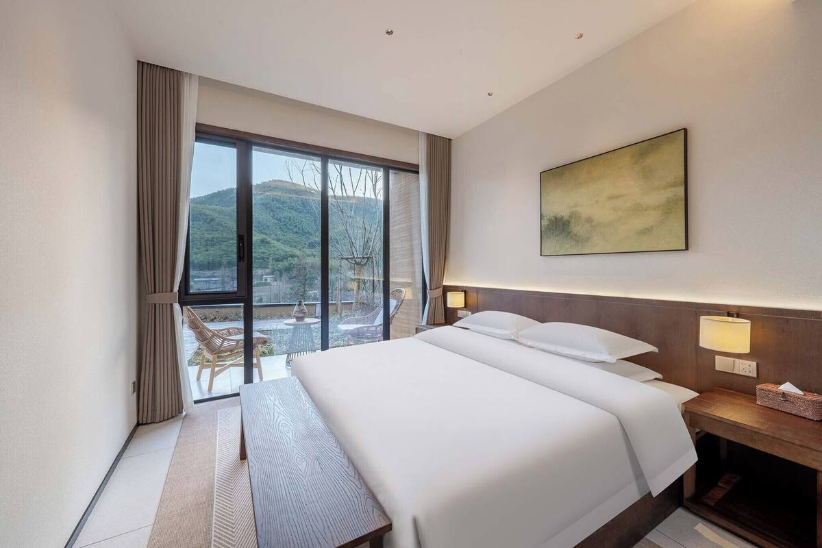 Habitación de hotel con una cama grande, un escritorio, una silla, una ventana con cortinas, una mesita de noche con lámpara y un cuadro en la pared.