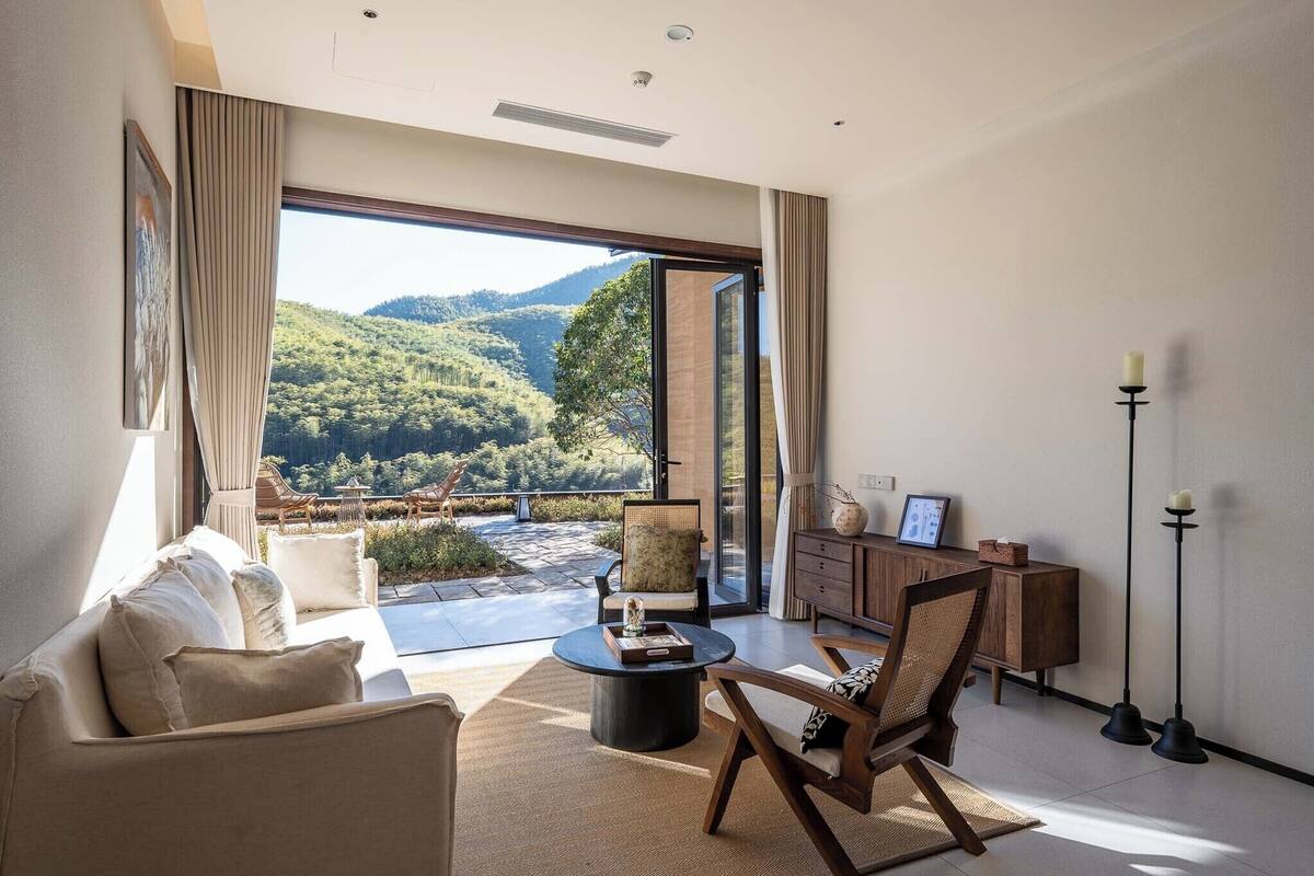 Una sala de estar moderna con un sofá, un sillón, una mesa de centro, un armario de madera y vistas a un paisaje montañoso a través de puertas de cristal grandes.