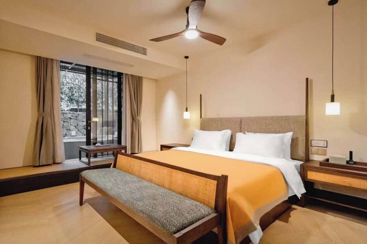 Habitación de hotel con cama, mesitas de noche, un banco, ventilador de techo y ventana con cortinas.