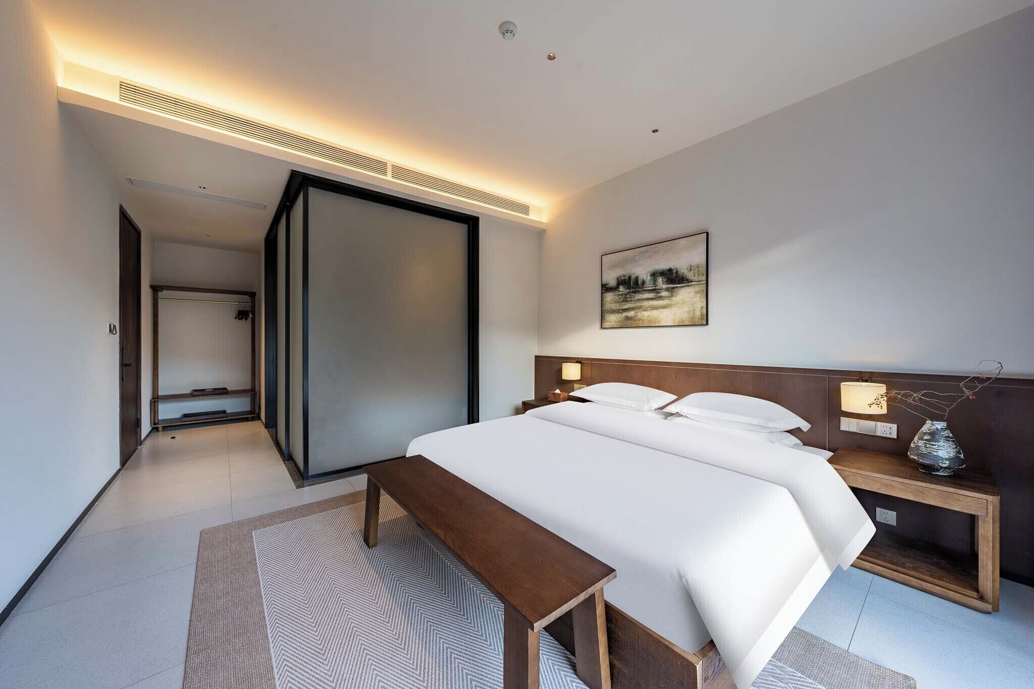 Una habitación de hotel moderna con una cama grande, mesitas de noche, un banco y un cuadro en la pared.