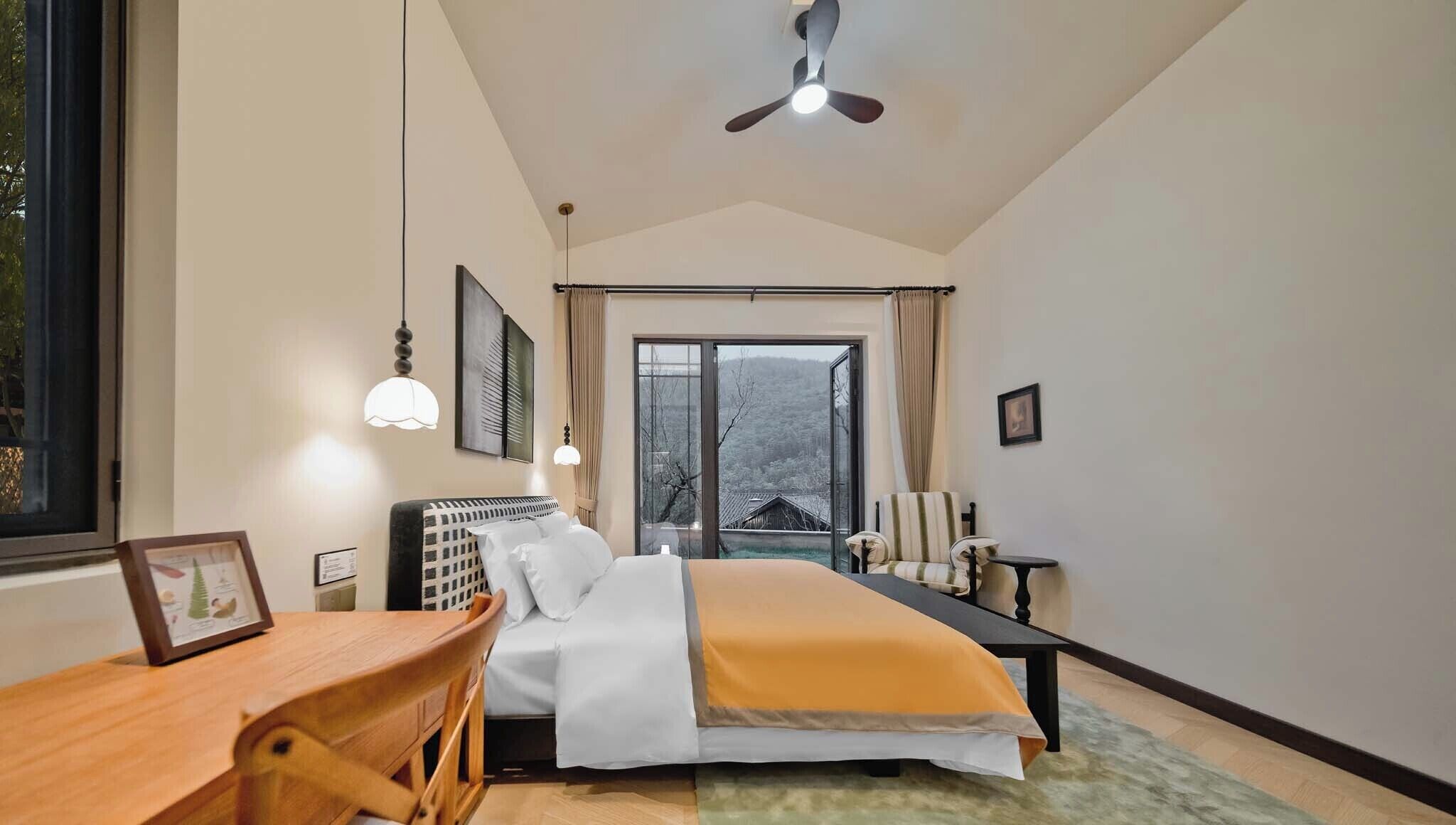 A bedroom with a bed, a desk, a chair, a ceiling fan, and a large window.