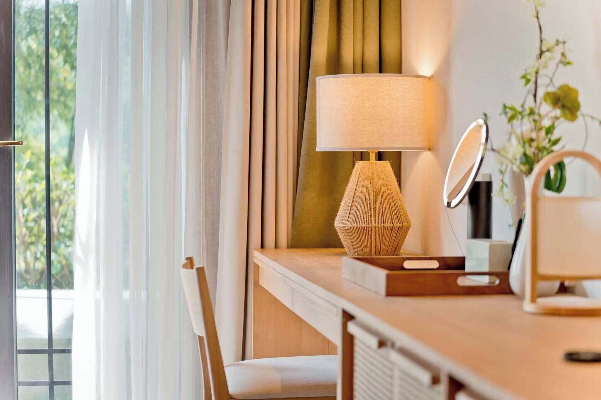 Habitación de hotel con escritorio de madera, lámpara, espejo y vistas a zonas verdes.