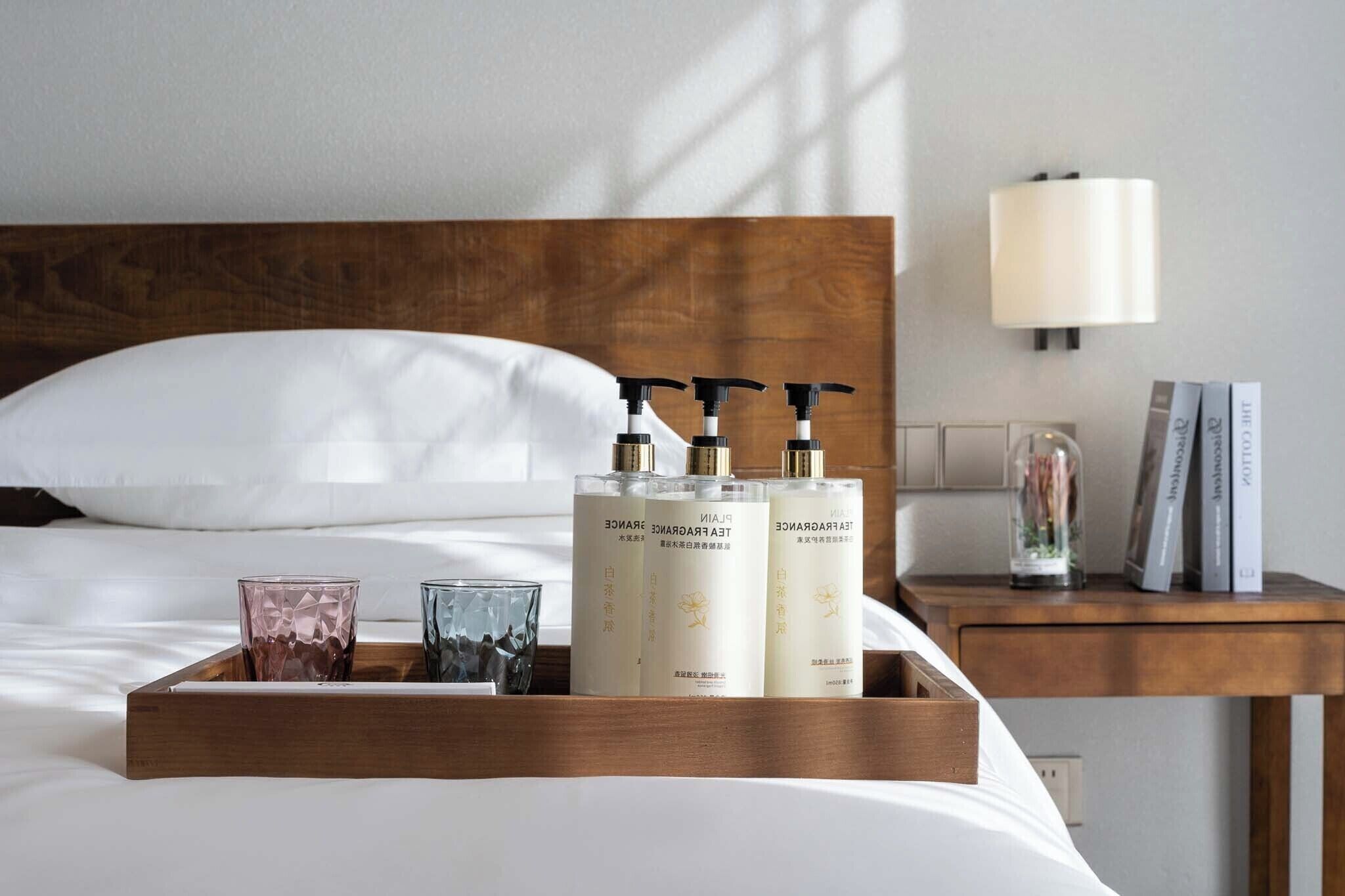 A neatly made bed with a wooden headboard, a bedside table with a lamp, and a tray with skincare products and glasses.
