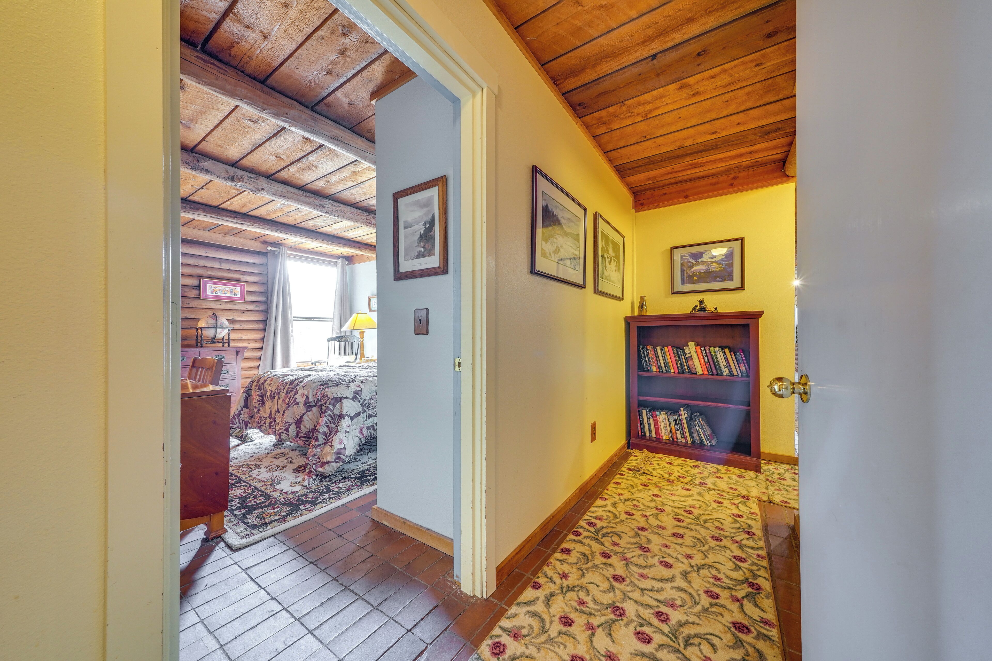A hallway with wooden ceiling beams, a patterned carpet, a bookshelf, and framed pictures on the wall.