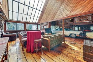 A spacious room with a high ceiling, wooden flooring, and a large window allowing natural light. There are multiple seating areas, including a sofa and a table with chairs.