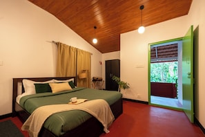 A bedroom with a wooden ceiling, a green door, a bed with a green bedspread, a bedside table with a lamp, and a view of greenery outside.
