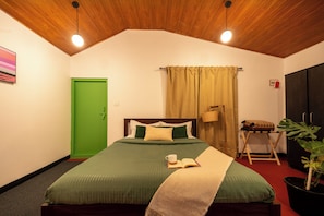 A bedroom with a green door, a bed with a green and beige bedspread, a bedside table with a lamp, and a potted plant.