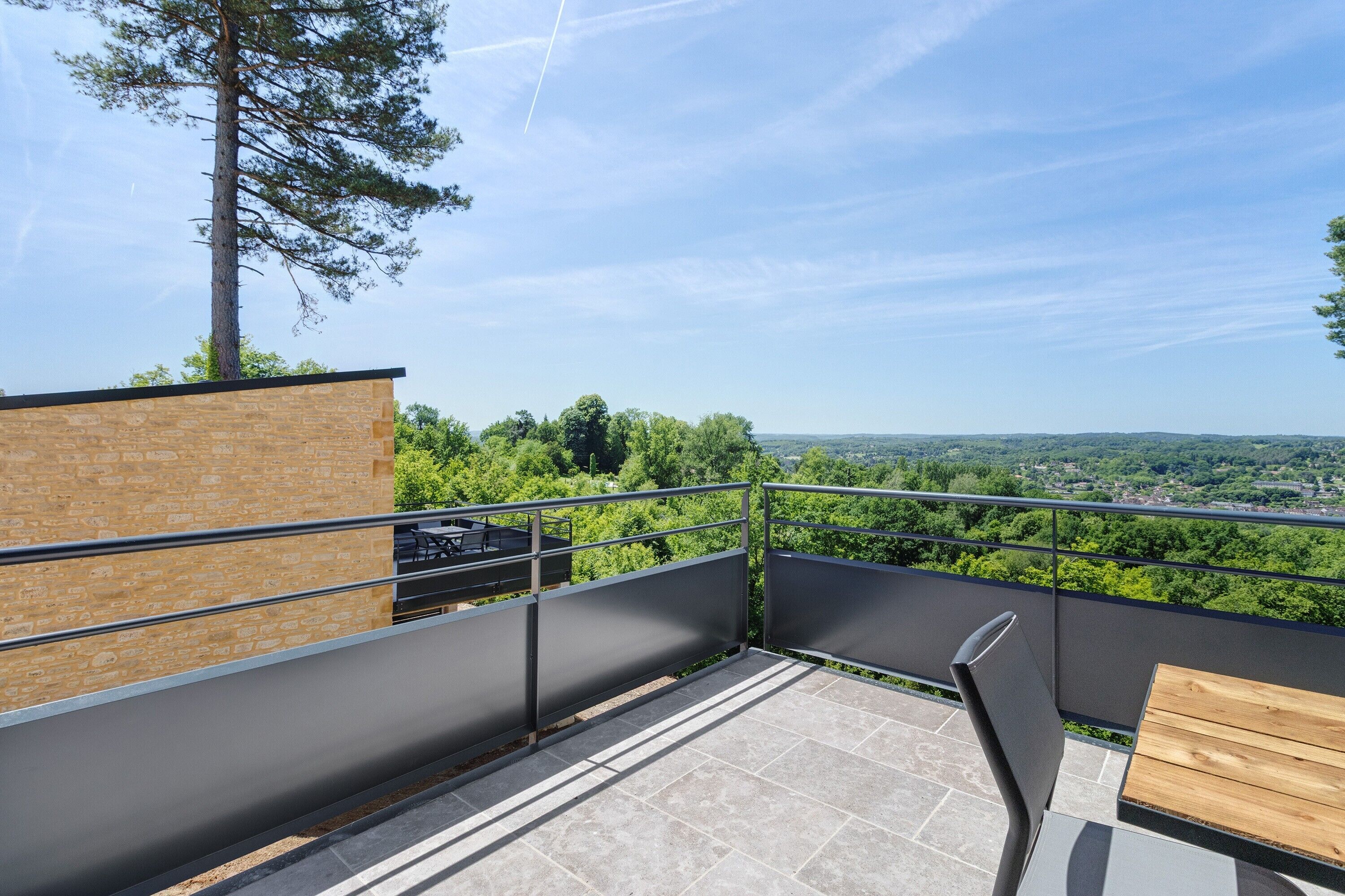 A balcony with a view of a forested landscape, a wooden table, and a metal railing.