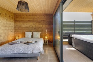 Quarto com cama, abajures, porta de vidro que dá para uma varanda com hidromassagem.
