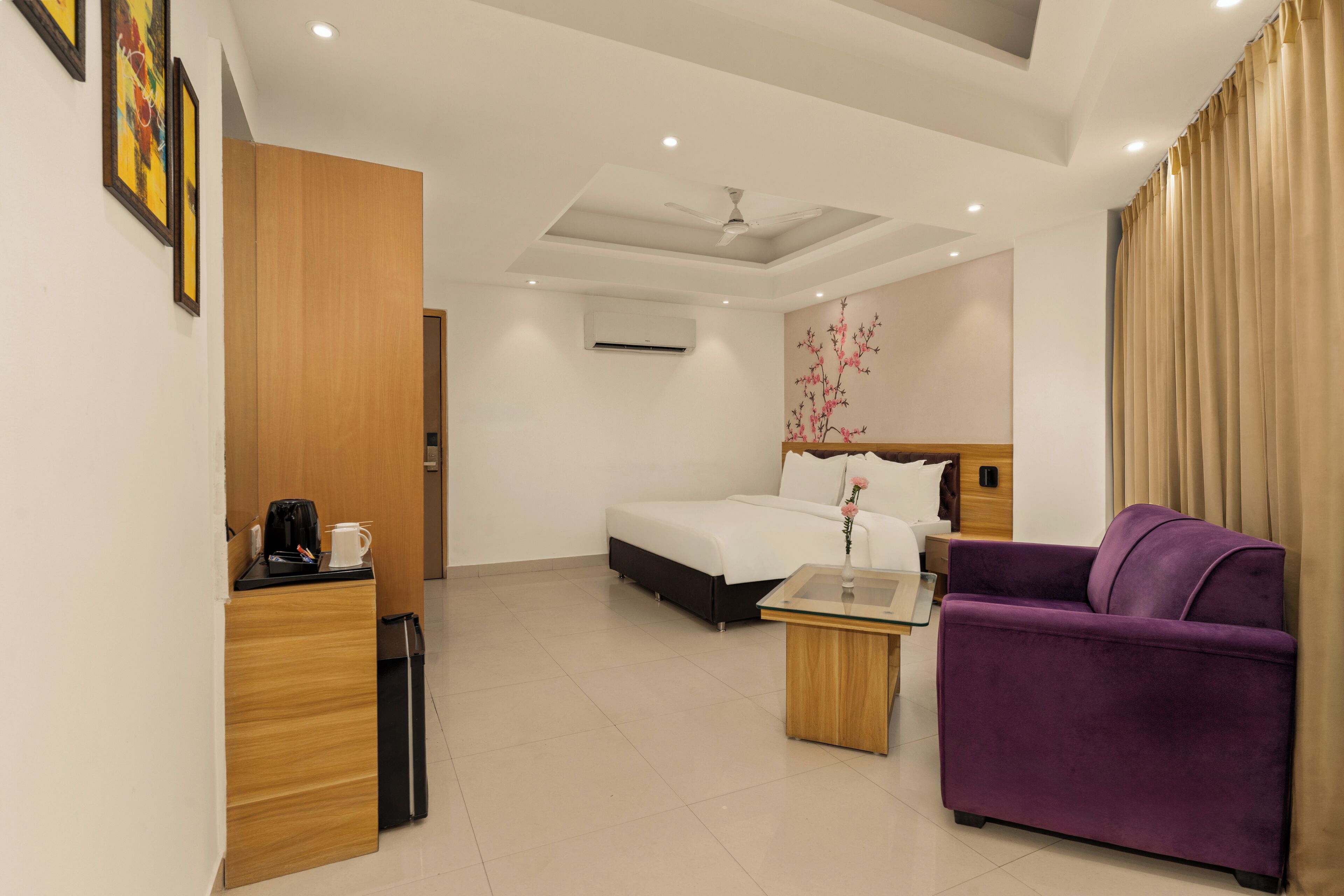 Habitación de hotel con cama, sofá púrpura, escritorio de madera con cafetera, una mesita y ventilador de techo.