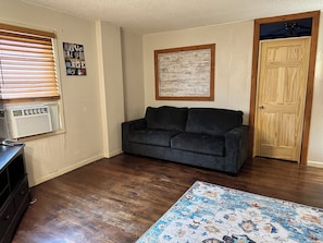 A living room with a dark grey sofa, a wooden door, a window with blinds, a wall-mounted air conditioner, and a patterned rug.