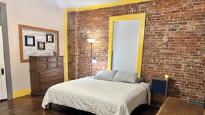 Een slaapkamer met een bakstenen muur, een bed met een witte spreidek, een dressoir, een lamp en een raam met jaloezieën.