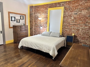 A bedroom with a brick wall, a bed with a white bedspread, a dresser, a lamp, and a window with blinds.