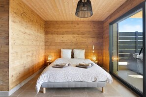 Quarto com teto de madeira, uma cama grande, duas luminárias de mesa e vista através de uma porta de vidro.
