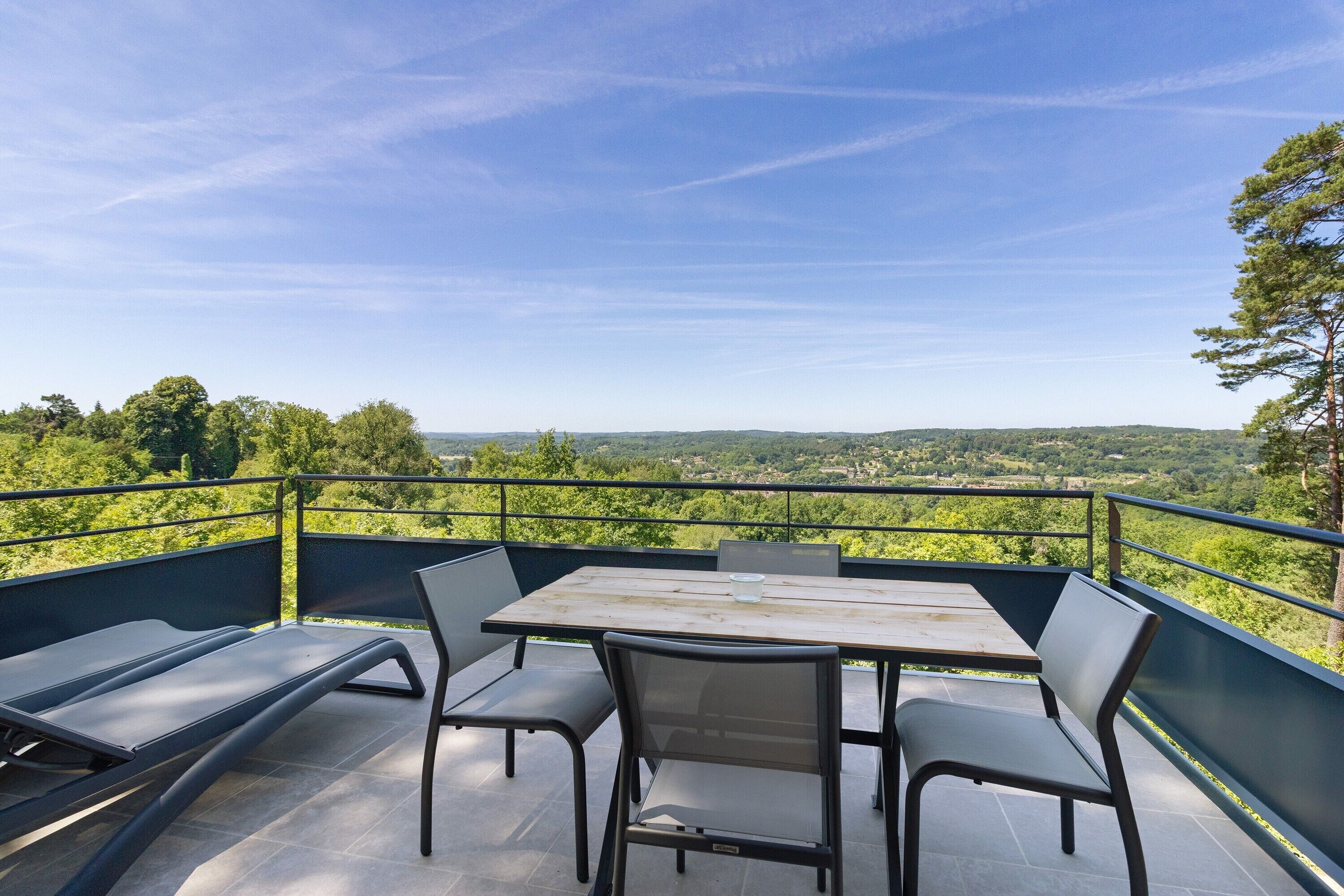 An outdoor terrace with a table and chairs, a lounge chair, and a view of a green landscape.
