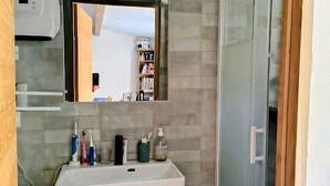 Un baño con lavamanos blanco, tocador de madera, un espejo y zona de ducha.