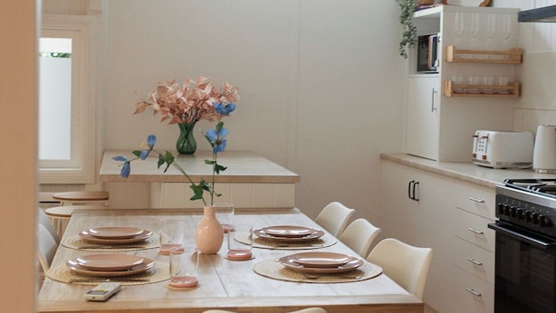 Spiseområde med et træbord dækket til fire personer, hvide stole, en vase med blomster, samt et køkken med hvide skabe og et vindue over vasken.