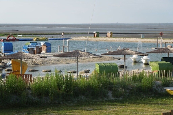 Ein Strand mit reetgedeckten Sonnenschirmen, einem Wasserspielbereich und einem Sandufer.