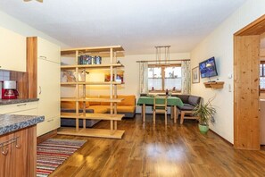 A compact living space with a kitchenette, a dining area with a table and chairs, a sofa, a television mounted on the wall, and a wooden bookshelf. - Holiday apartment in Waidring with Wi-Fi (Waidring)