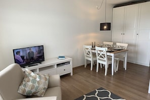 Ein Wohnzimmer mit Fernseher, weißen Möbeln, einem Esstisch und einem Kleiderschrank.