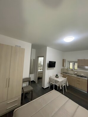 Et lille hotelværelse med seng, spisebord med stole, tekøkken og et tv ophængt på væggen.